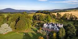 Aerial view of a large white mansion, the Douneside House & Health Club surrounded by lush green trees and rolling hills.