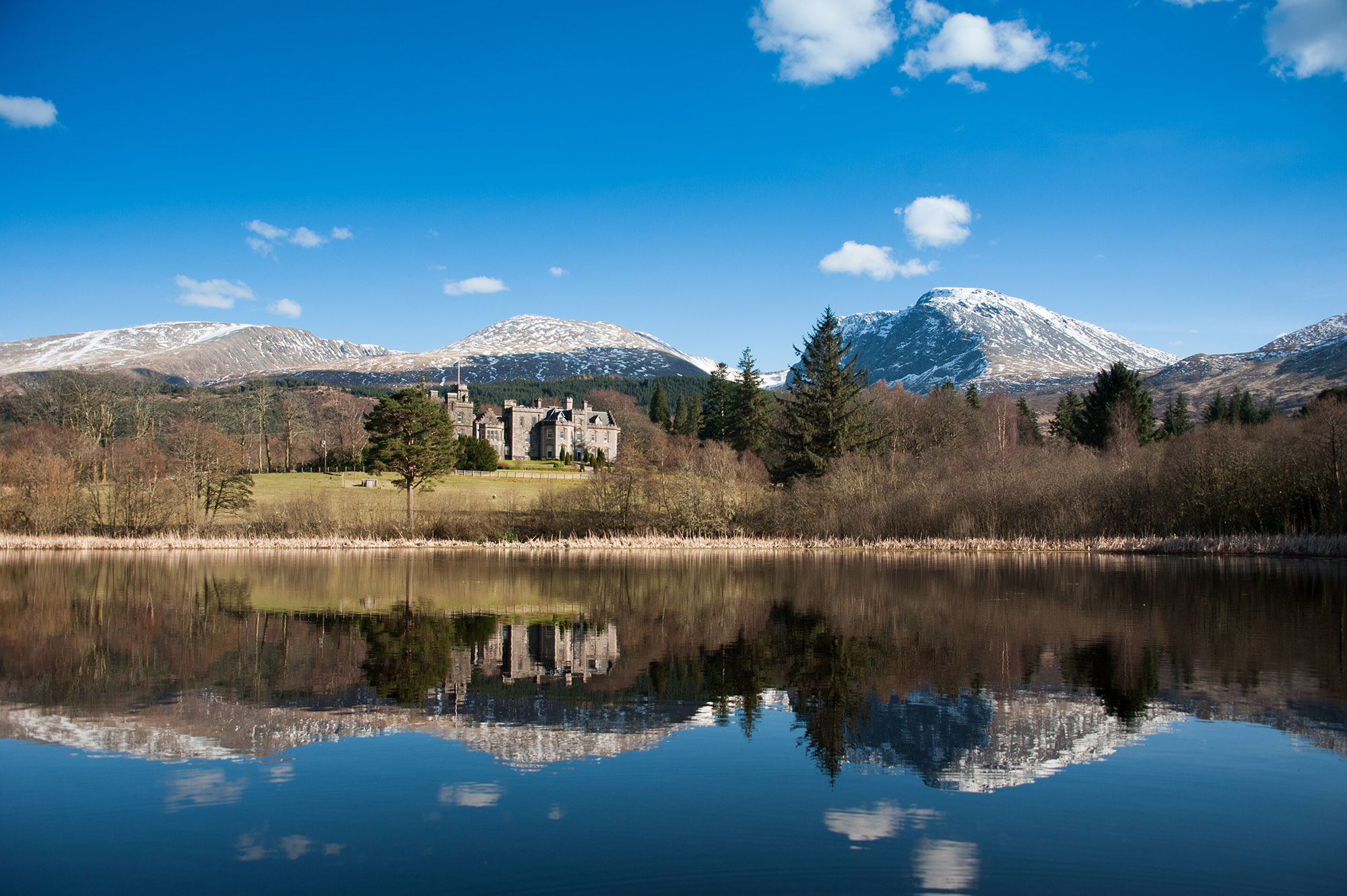 A castle beside a calm lake, with snowy mountains in the background.