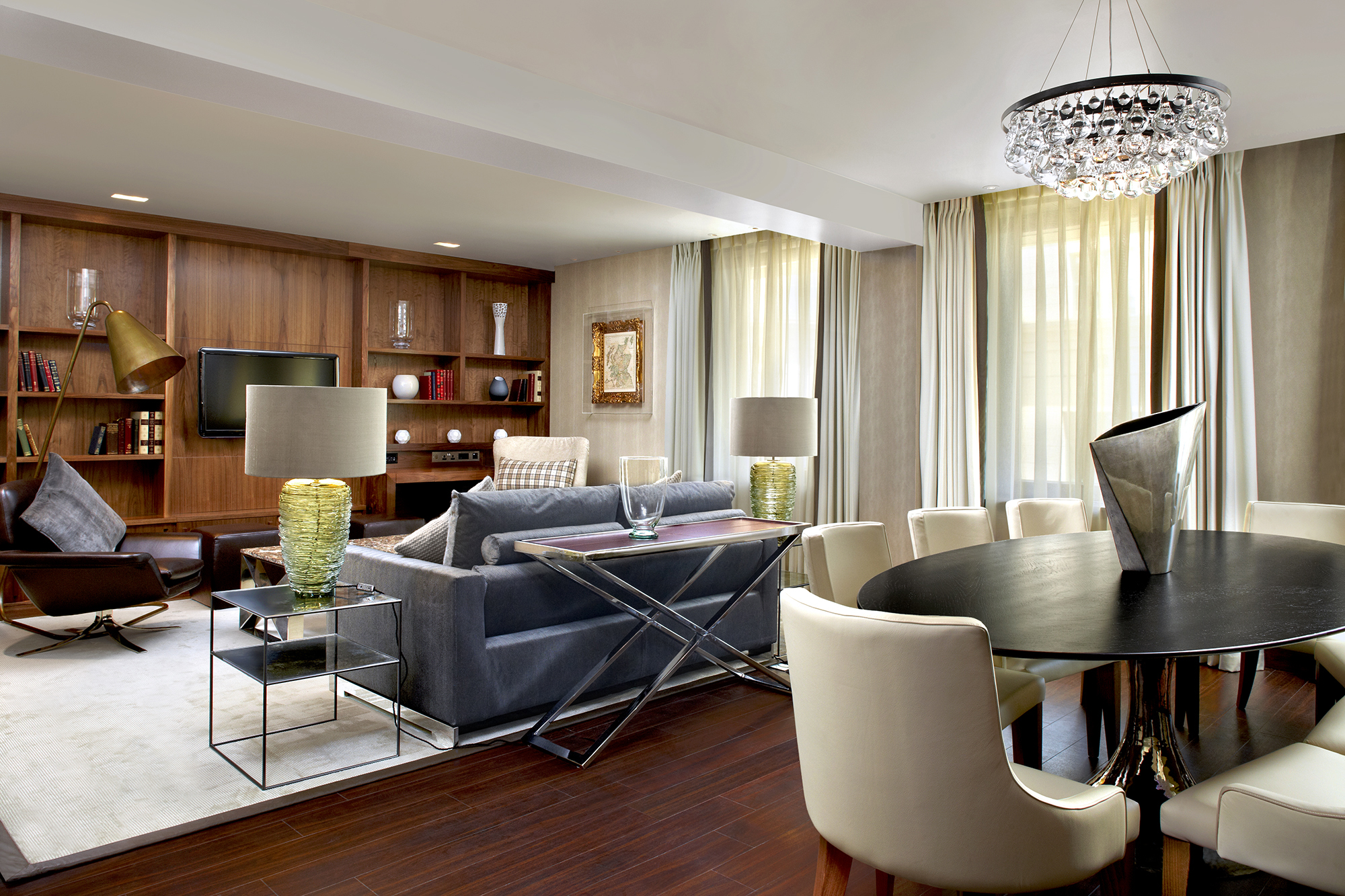 Modern suite with a round dining table, velvet chairs, and a cosy lounge under a crystal chandelier.