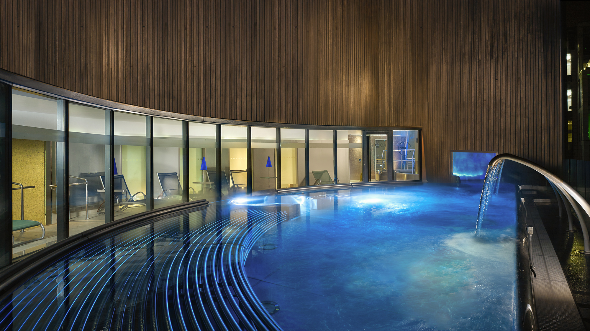 Modern spa hydro pool with curved design and waterfall feature, glowing in blue light.
