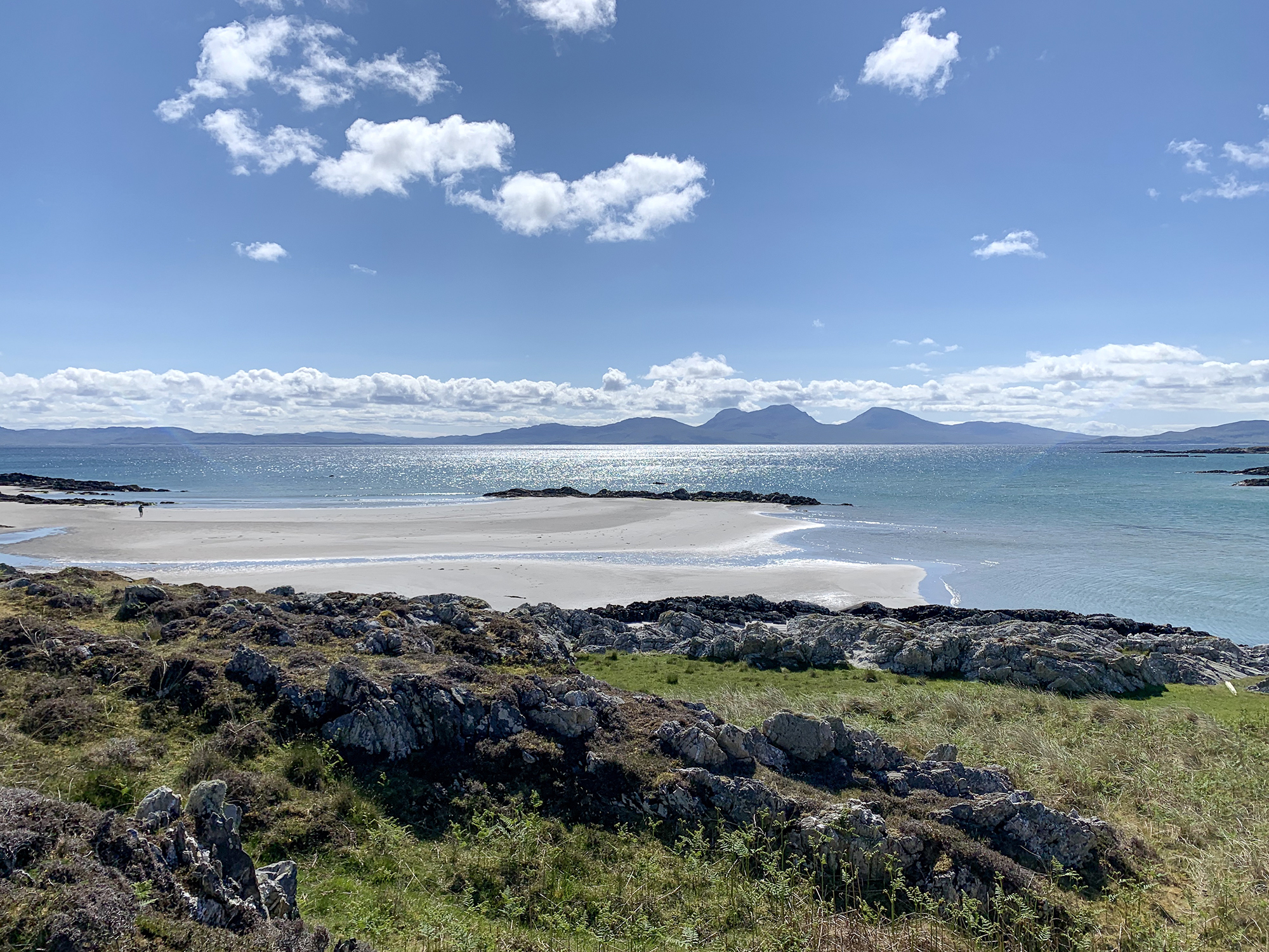 Beach view with sandy shores, rocky terrain, and distant mountains under a bright sky on the Isle of Jura.