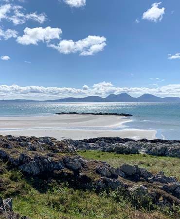 Beach view with sandy shores, rocky terrain, and distant mountains under a bright sky on the Isle of Jura.