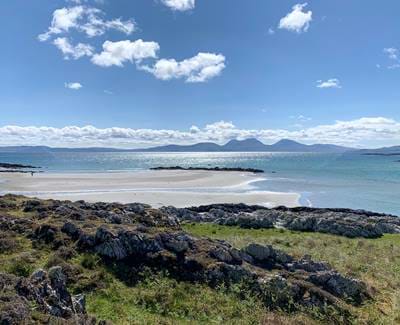 Beach view with sandy shores, rocky terrain, and distant mountains under a bright sky on the Isle of Jura.