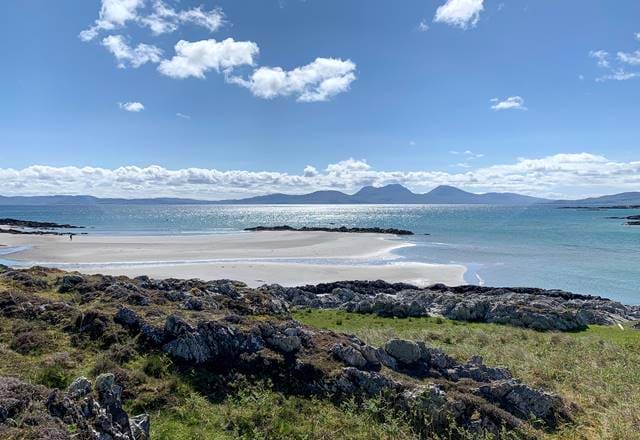 Beach view with sandy shores, rocky terrain, and distant mountains under a bright sky on the Isle of Jura.