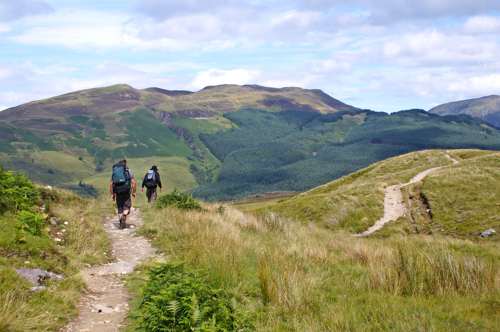 Two hikers with backpacks walk a grassy trail toward forested hills under a partly cloudy sky in Scotland.