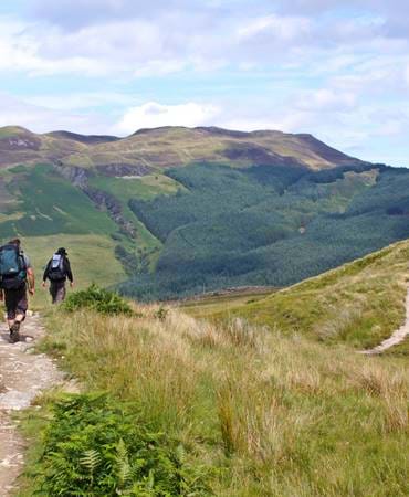 Two hikers with backpacks walk a grassy trail toward forested hills under a partly cloudy sky in Scotland.