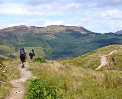 Two hikers with backpacks walk a grassy trail toward forested hills under a partly cloudy sky in Scotland.