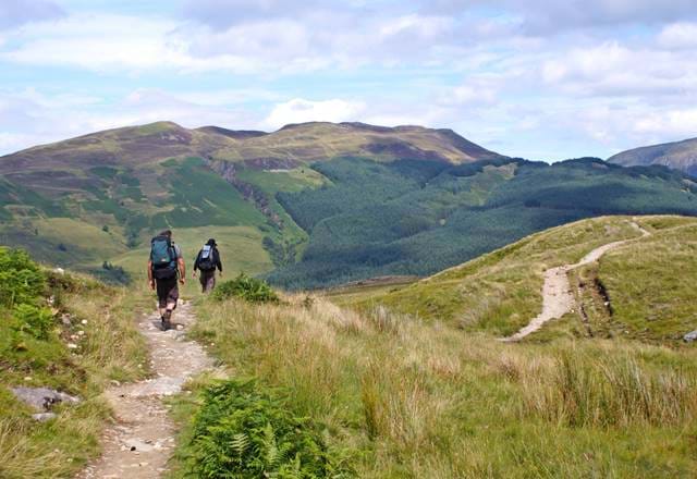 Two hikers with backpacks walk a grassy trail toward forested hills under a partly cloudy sky in Scotland.