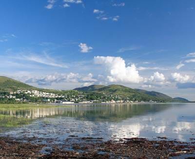 Scenic coastal Scottish seaside town with white houses nestled between green hills and reflective water.