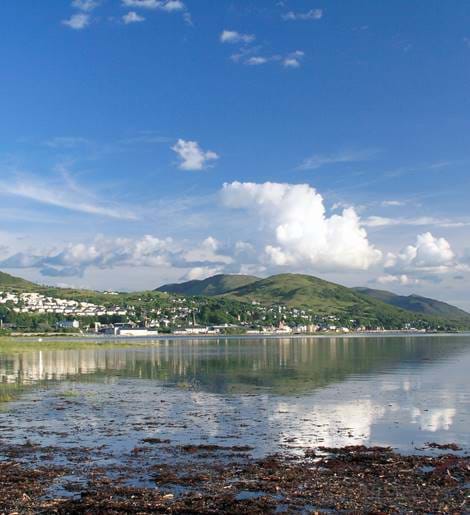 Scenic coastal Scottish seaside town with white houses nestled between green hills and reflective water.
