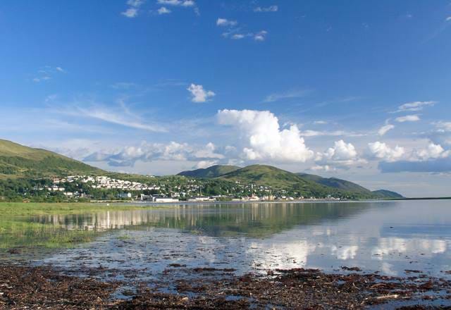 Scenic coastal Scottish seaside town with white houses nestled between green hills and reflective water.