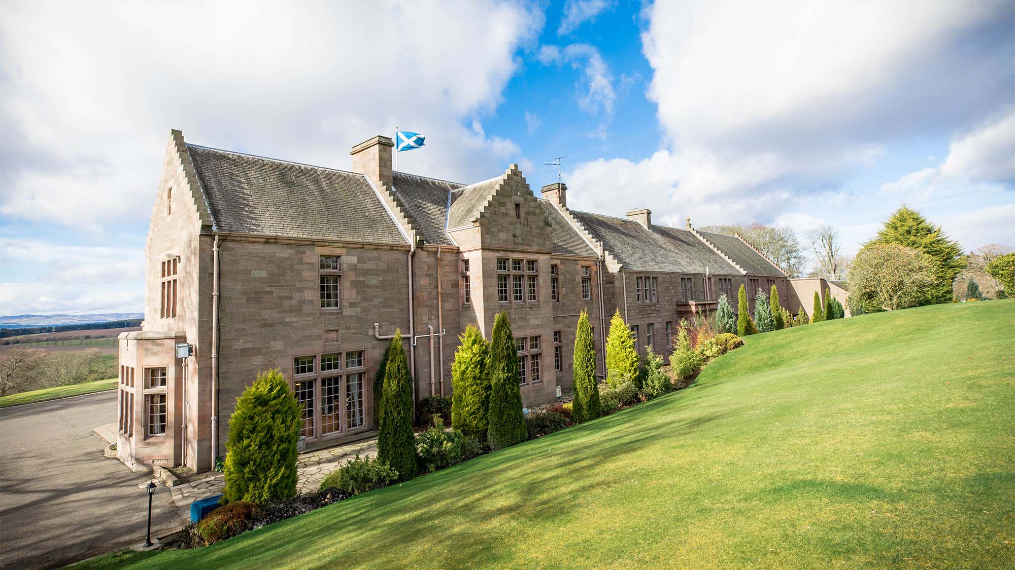 The Scottish stone house of the Murrayshall Hotel with a flag flying, nestled within a manicured lawn.