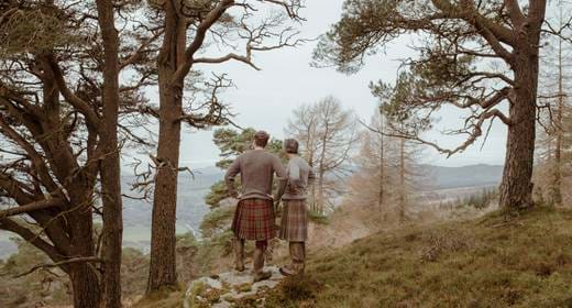 Two people wearing Highland Outfitters kilts, standing side by side in a forested landscape, gazing at the view.
